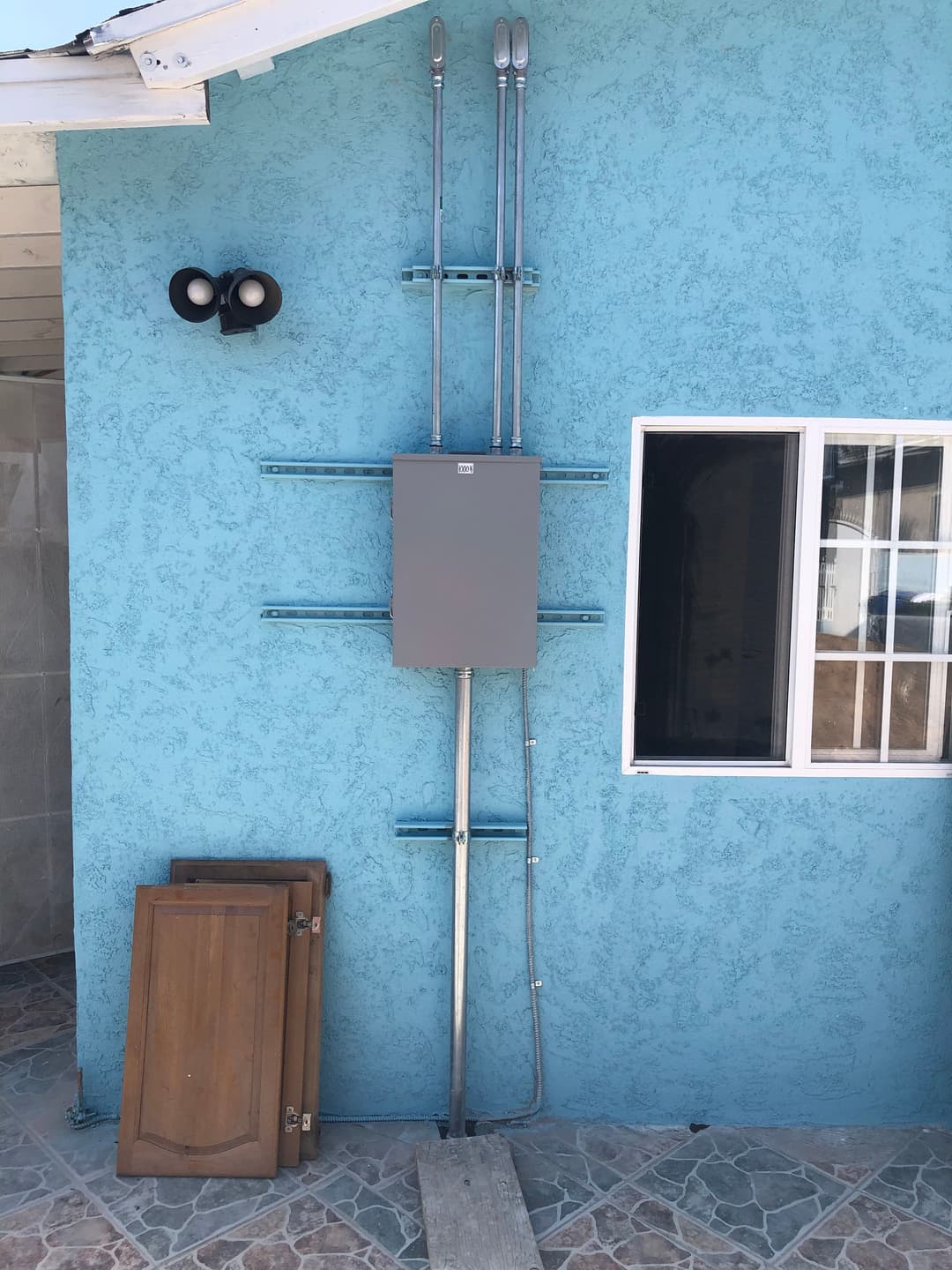 Electrical panel mounted on a blue exterior wall with metal conduits and wooden doors beside it.