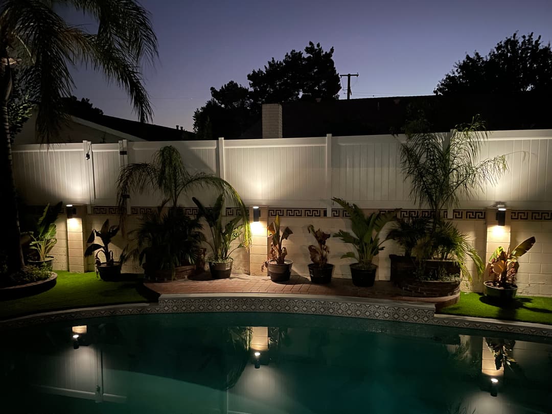 Illuminated backyard pool at night with plants and a white fence.