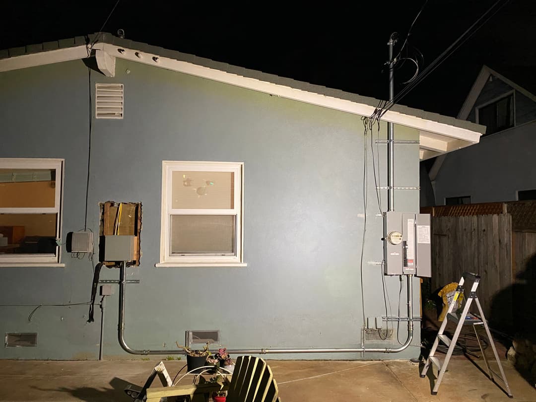 Evening exterior view of a green house with electrical panel and ladder in the foreground.