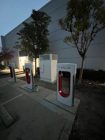 Tesla Superchargers in an outdoor charging station at dusk, alongside trees and a wall.